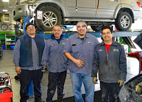 Four auto mechanics standing in auto shop with car on a lift behind them.